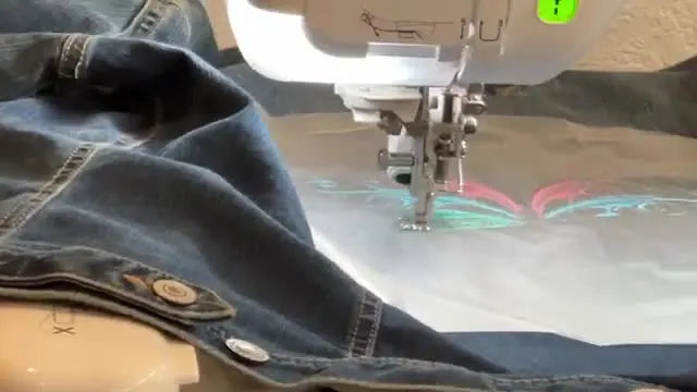 Extreme close-up of the Brother Dream Machine stitching pink thread details into a butterfly design on denim with a water-soluble topper on top.