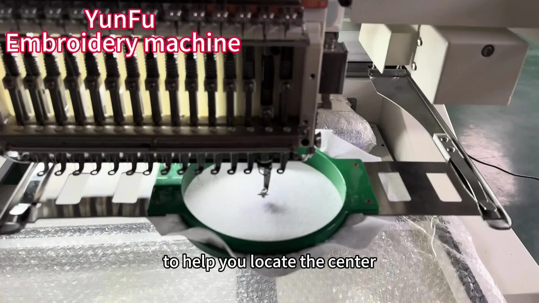 A close-up shows an operator pressing the needle bar down to verify the needle tip lands exactly on the hand-drawn center crosshair inside the T-shirt frame.