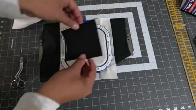 Hands press a square of Velcro onto the back side of the hooped patch before the final tack-down run stitch.
