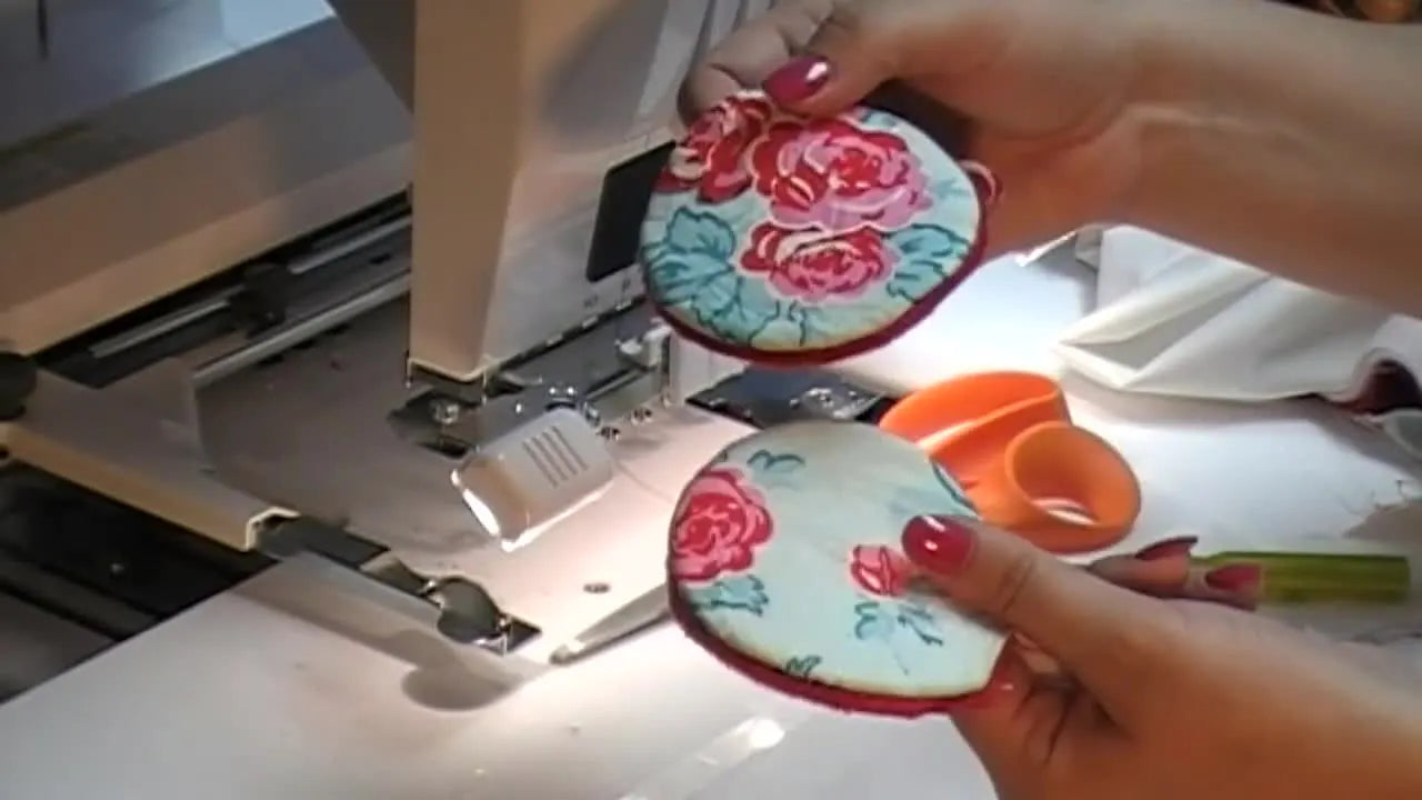 The host holds up two finished floral in-the-hoop breast pads, showing the clean edges and professional shape after turning and stitching the opening closed.